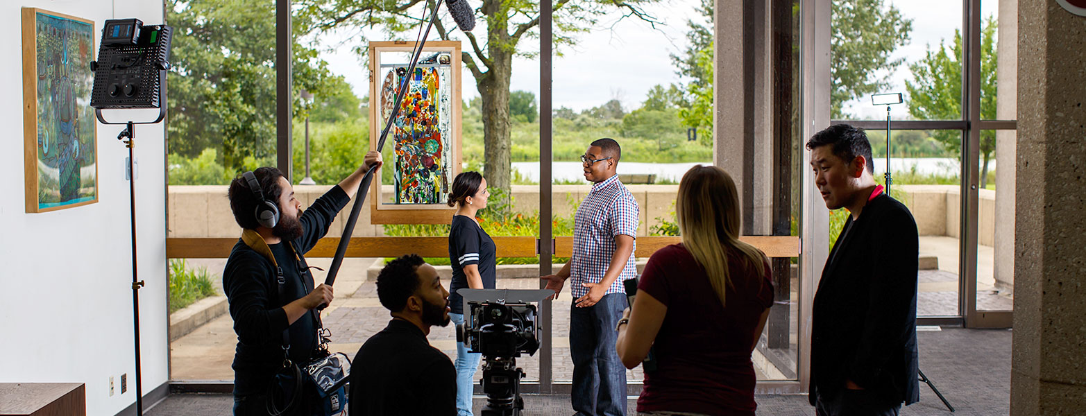 Students with digital video camera, lights, and boom microphone filming a scene with two actors.