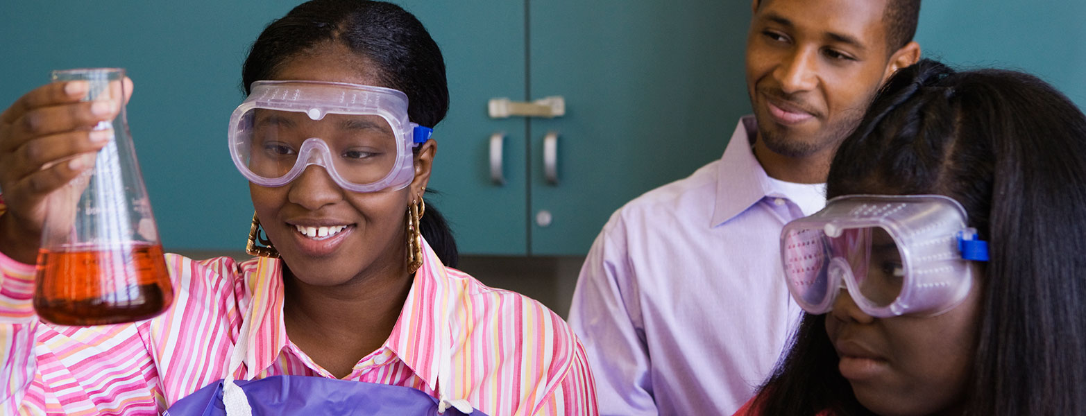 Students with goggles in chemistry lab looking at red liquid in a beaker.