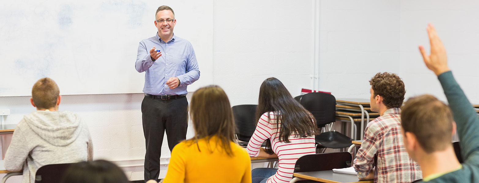 Male teacher in front of whiteboard calling on a student in class.