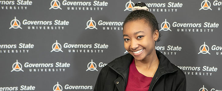 Student smiling while standing in front of back drop with a repeated Governors State logo Student smiling while standing in front of back drop with a repeated Governors State logo