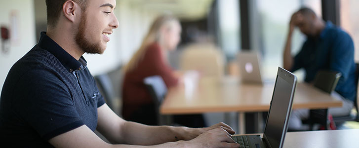 Student typing on laptop at a hallway desk on campus Student typing on laptop at a hallway desk on campus