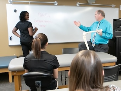 Professor holding model skeleton leg while talking to a student writing on a dry erase board in front of class