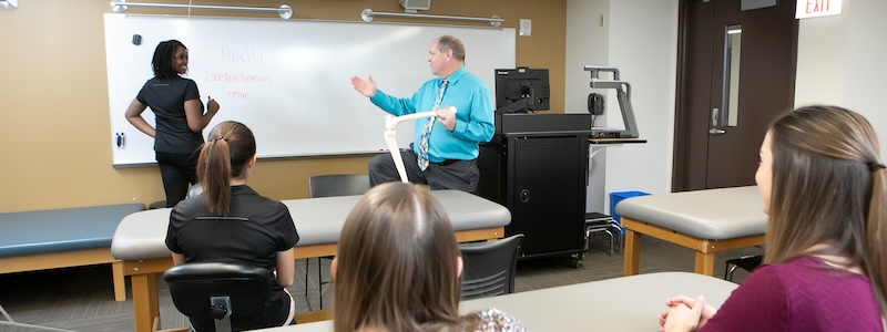 Professor holding model skeleton leg while talking to a student writing on a dry erase board in front of class