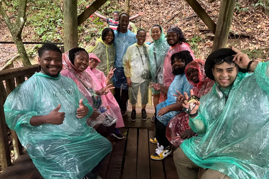 A group of people wearing colorful rain ponchos gather on a wooden platform in a lush, forested area. They are smiling and gesturing with thumbs up, indicating a cheerful mood. The setting appears to be a nature trail or park. Among other alumni, Marquis Parks, Alexis Berry and Jocelyn Bell are pictured.