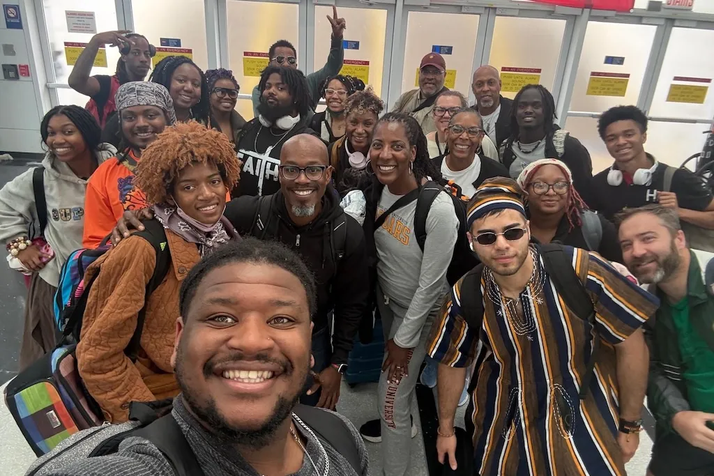 A group of people stands together, smiling for a photo indoors. Some are holding bags with colorful designs. The background includes a row of red and white signs labeled "Stairs." The atmosphere is cheerful and relaxed. Among other alumni, Marquis Parks, Alexis Berry, Jocelyn Bell and Inala Bruce-Rose are pictured.