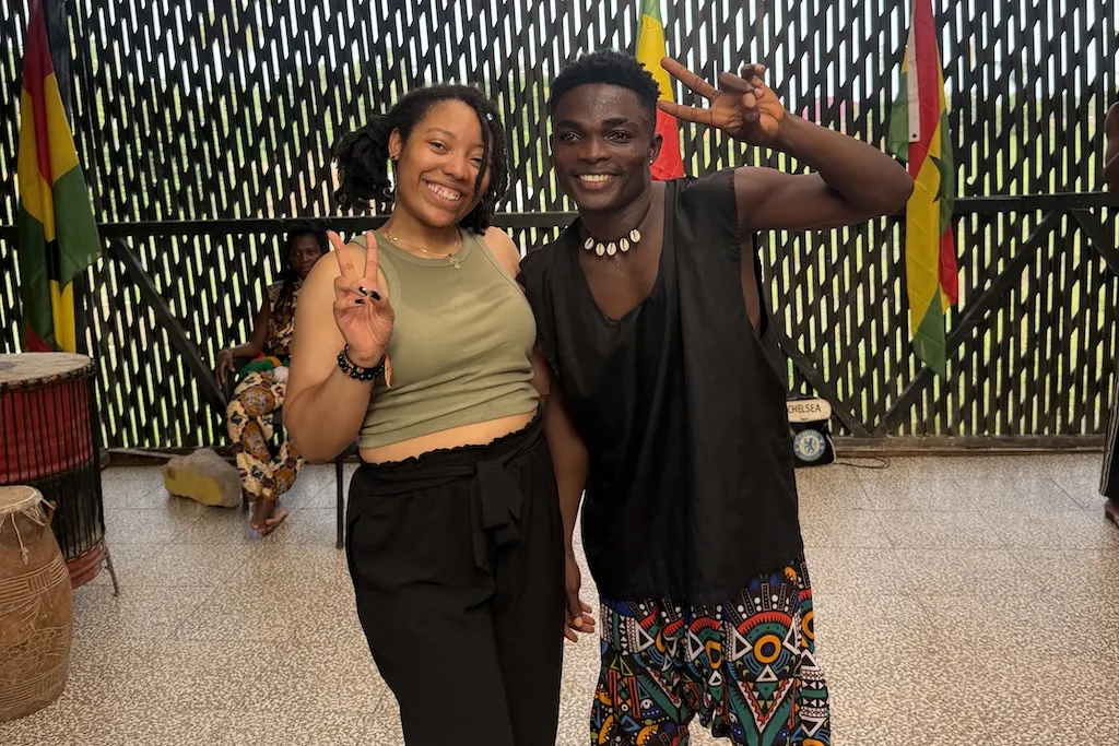 Two people smiling and making peace signs. They are standing in front of a fence with colorful flags. The person on the right is wearing a black outfit with colorful, patterned pants. The setting appears casual and relaxed. GovState alumna, Inala Bruce-Rose, is pictured on the left.