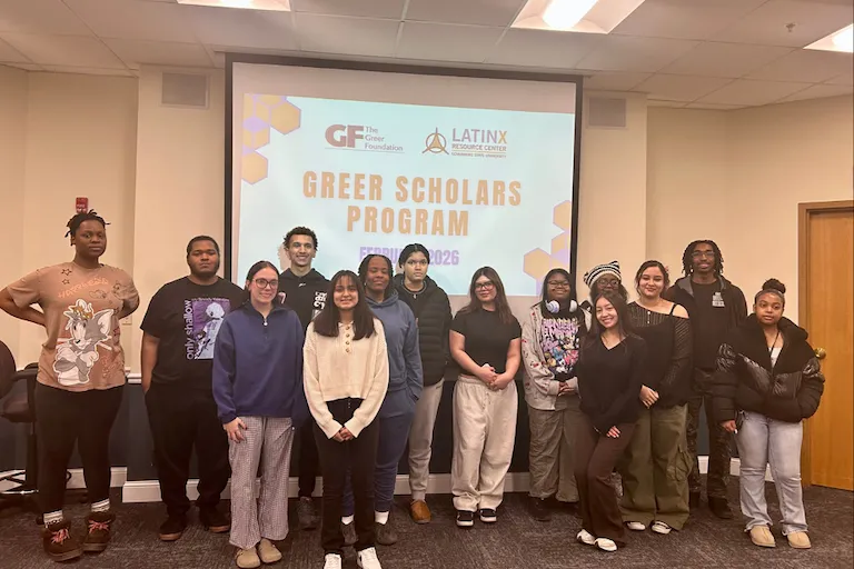 A group of students in front of a screen displaying "GREER SCHOLARS PROGRAM."