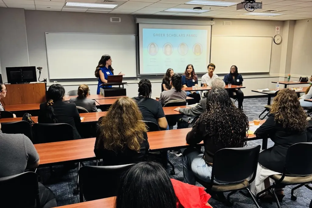 Panel discussion with a speaker at a podium, audience, and projector screen in a classroom.