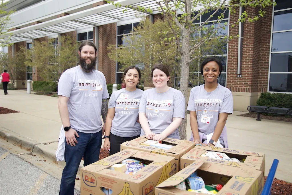 Four smiling volunteers in gray shirts with boxes of food at an outdoor food drive.