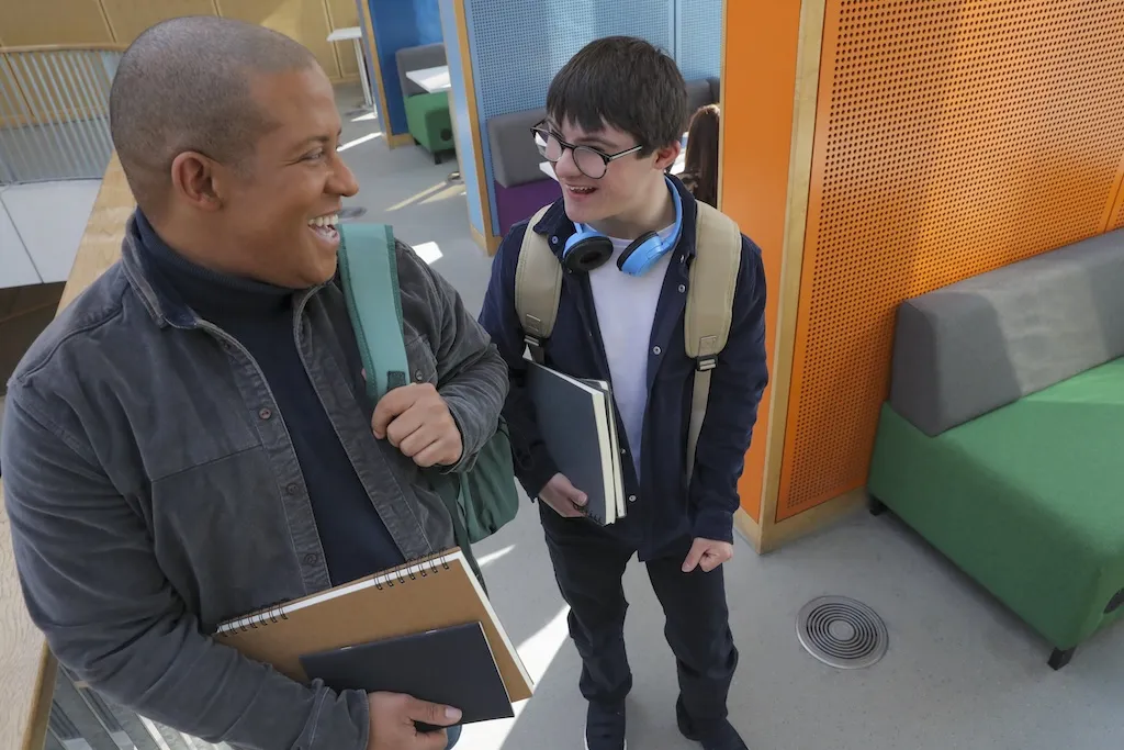 Two smiling students chat while carrying books in a modern, colorful building.