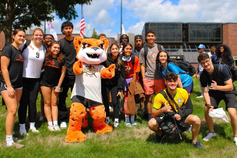 Diverse group of smiling students and an orange tiger mascot on a sunny campus lawn.