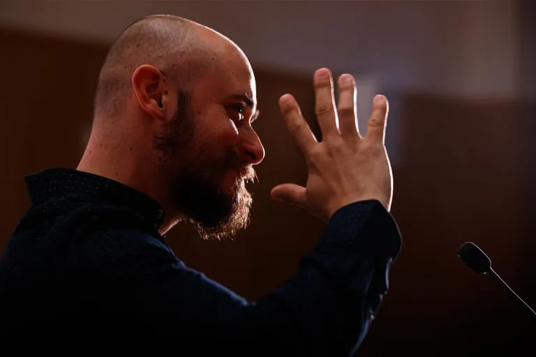 Bald, bearded man gestures with raised hand while speaking at a microphone.