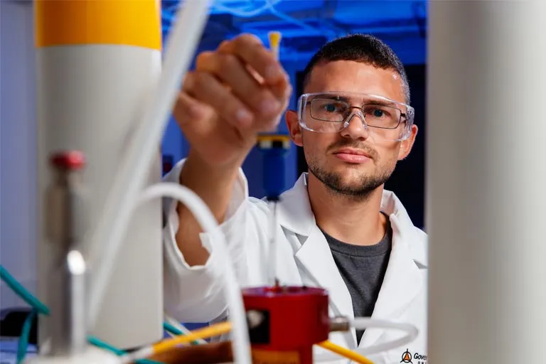 Student in safety glasses and lab coat holds a small yellow object.