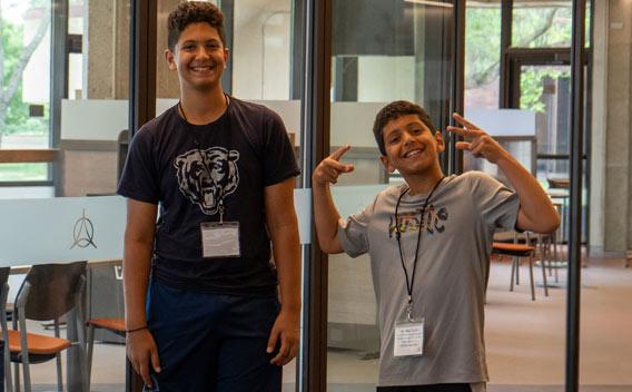 Two smiling young males with lanyards, one making peace signs, in a modern building.