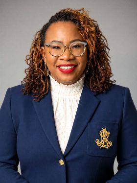 Professional headshot of a smiling woman with curly hair, glasses, and a navy blazer with a gold emblem.