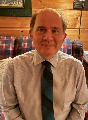 Man in a striped shirt and blue-green tie, smiling against a wooden wall and plaid sofa.