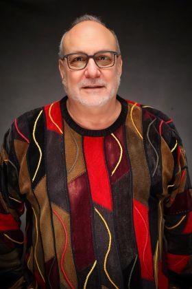 Man with glasses and beard, wearing a vibrant patterned sweater, smiling.