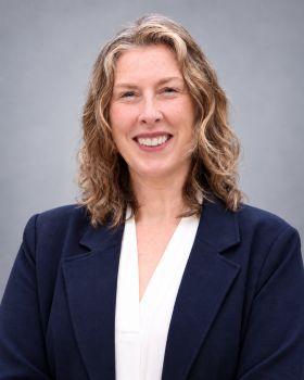 Smiling woman with wavy brown hair, wearing a dark blue blazer.