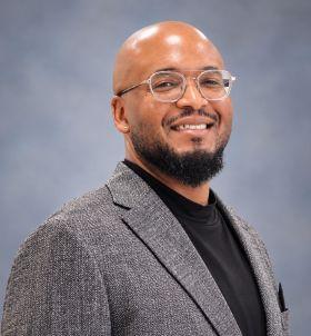 Smiling man with glasses, beard, gray blazer, and black shirt.
