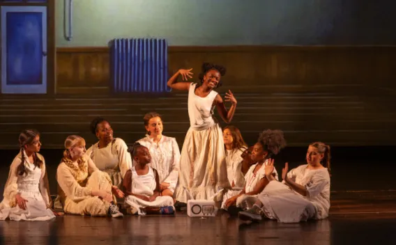 A group of performers in light-colored costumes sits onstage around a standing performer striking a dramatic pose