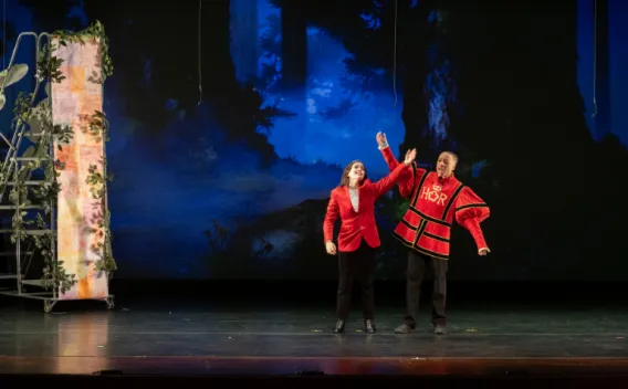 Two performers stand onstage in front of a dark forest backdrop, one in a red jacket and one in a red theatrical costume, raising an arm upward