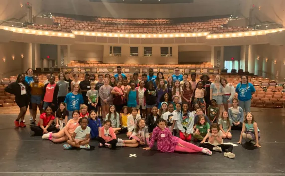 A large group of children and adults gather for a group photo on a theater stage with empty audience seating in the background