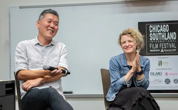 Two people seated in discussion at a conference or panel event in front of a whiteboard.