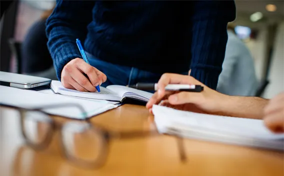 Hands writing in a notebook and reviewing papers on a wooden table.