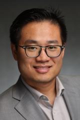 Professional headshot of an Asian man with glasses, smiling in a gray suit.