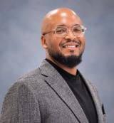 Smiling man with glasses, beard, gray blazer, and black shirt.