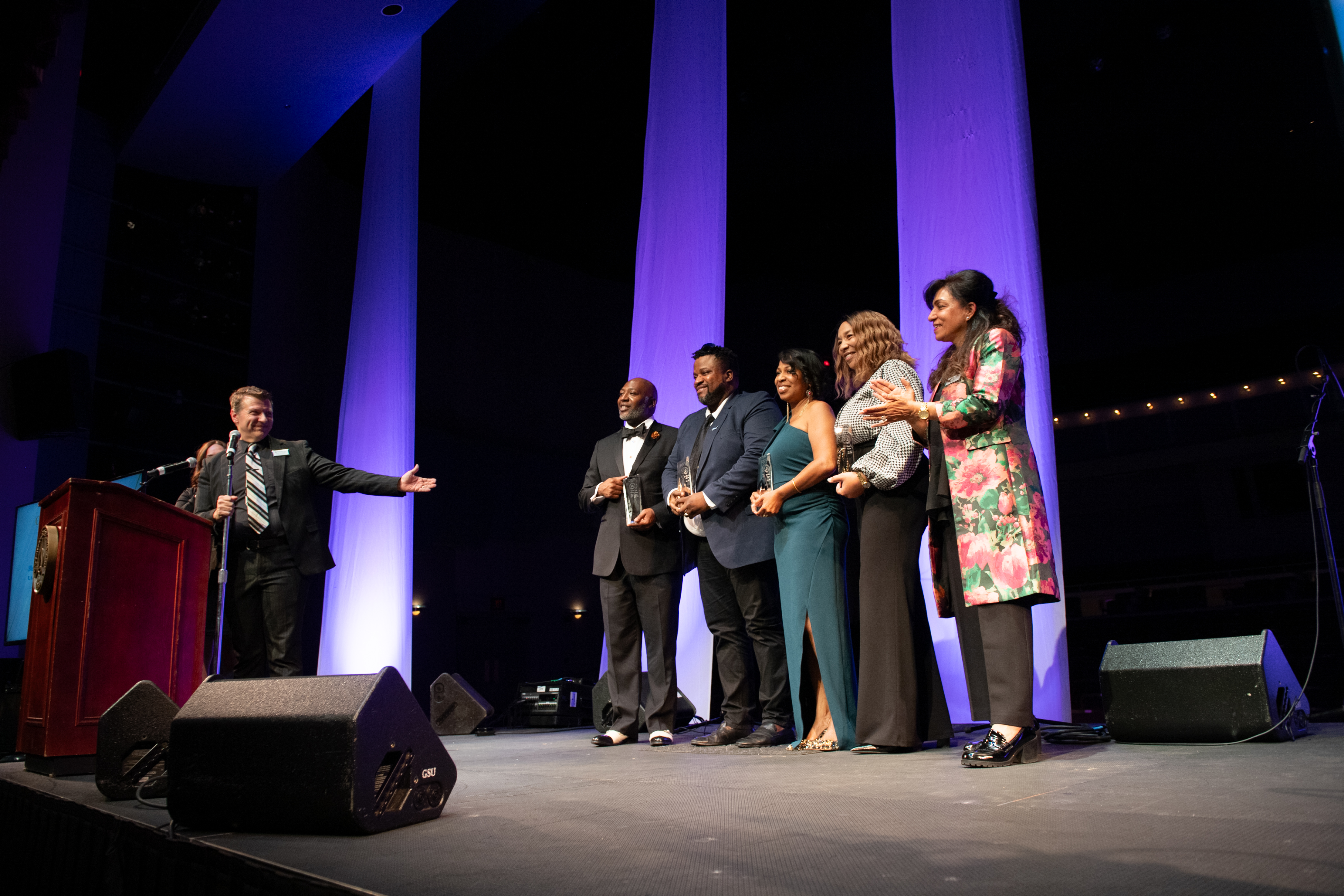 An event host presents a group of smiling awardees on stage during the 2025 Distinguished Alumni Reception at Governors State University An event host presents a group of smiling awardees on stage during the 2025 Distinguished Alumni Reception at Governors State University